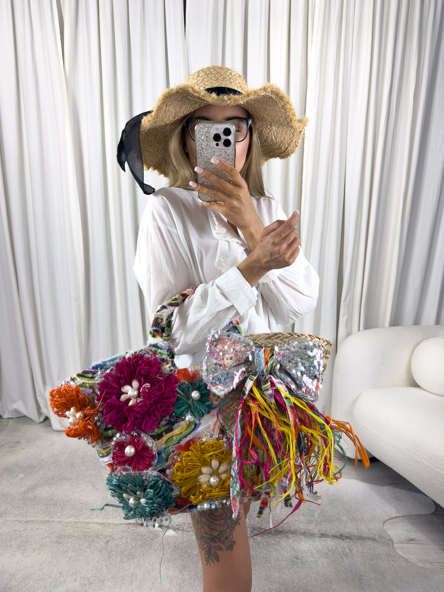 Letnia torebka koszyk z kolorowymi kwiatami, kokardą i frędzlami w stylu boho, trzymana przez kobietę w słomkowym kapeluszu. Zdjęcie produktowe sklepu internetowego z odzieżą damską.
