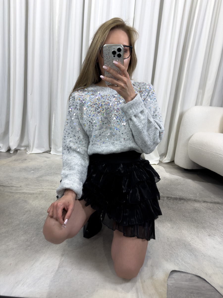 Sweter Sequins jasny szary z okrągłym dekoltem i cekinami w srebrnym odcieniu, zestawiony z czarną mini z falbanami – efektowna i elegancka stylizacja na zimowe okazje.