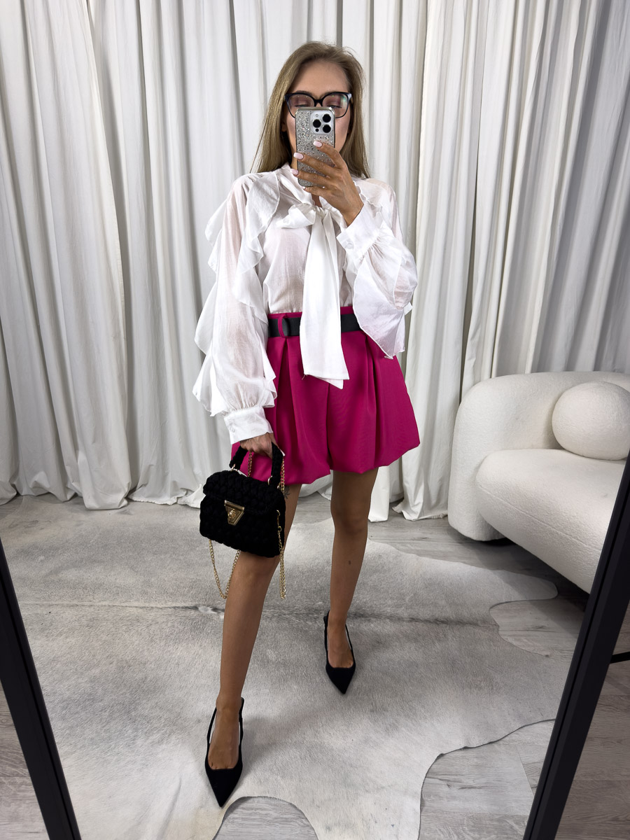 Zdjęcie produktowe sklepu internetowego z odzieżą damską – modelka ubrana w koszule Bow w kolorze białym marki Miho’s, spódnico-spodenki w kolorze fuksji, trzymająca w ręce małą, czarną torebkę