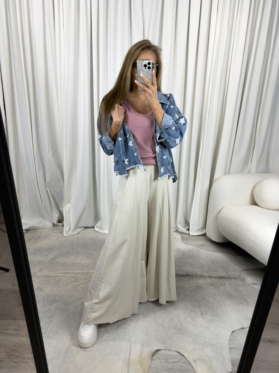 Modelka prezentuje luźne spodnie Baggy Wiya w beżowym kolorze, z szerokimi nogawkami i wysokim stanem, zestawione z różowym topem i jeansową kurtką z gwiazdkami, w stylizacji casualowej na tle jasnych zasłon.