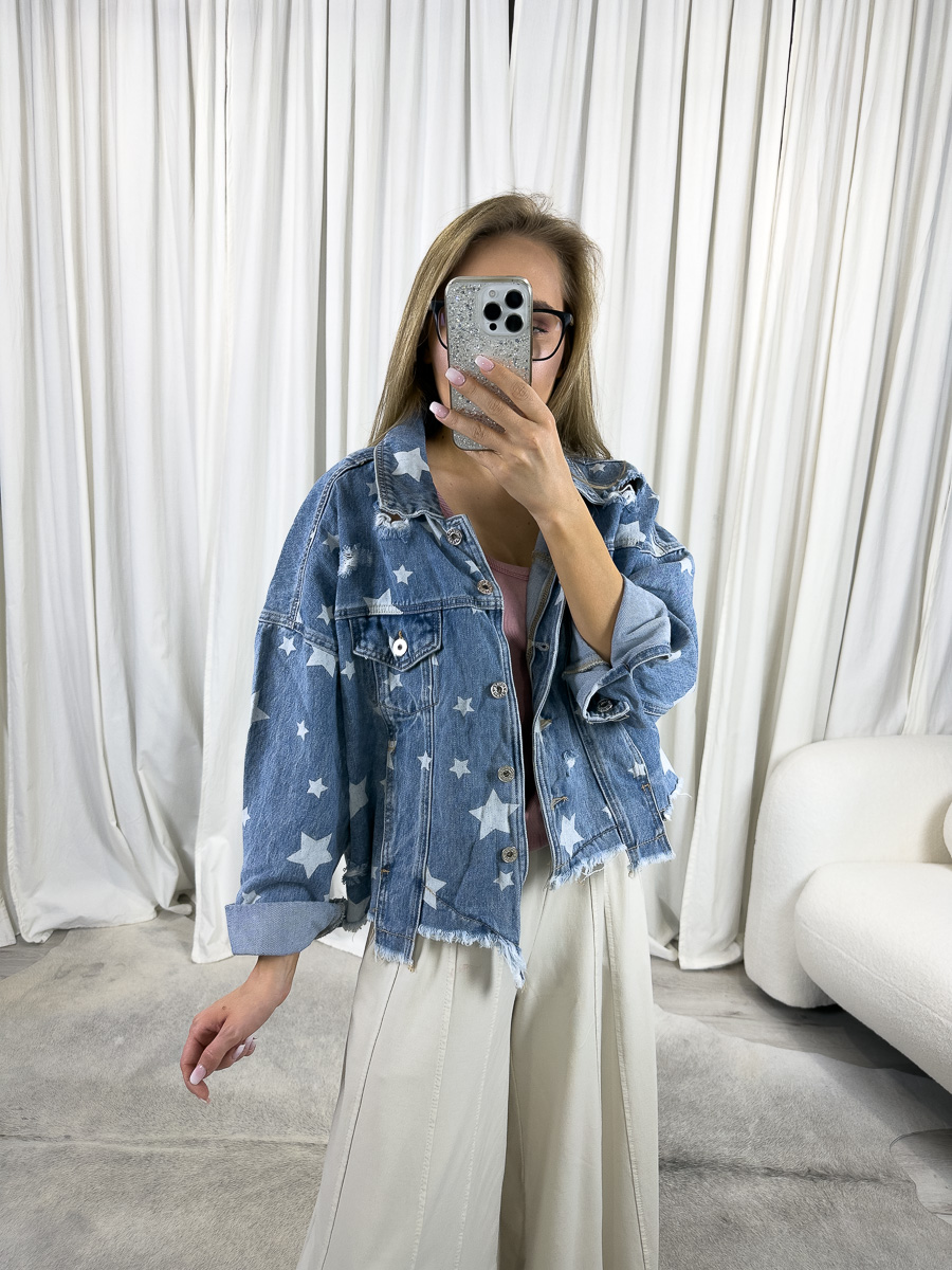 Modelka prezentuje kurtkę jeansową Stars z nadrukiem białych gwiazdek, o oversize'owym kroju i postrzępionym dole, zestawioną z jasnymi, szerokimi spodniami i różowym topem. Stylizacja casual na tle białych zasłon