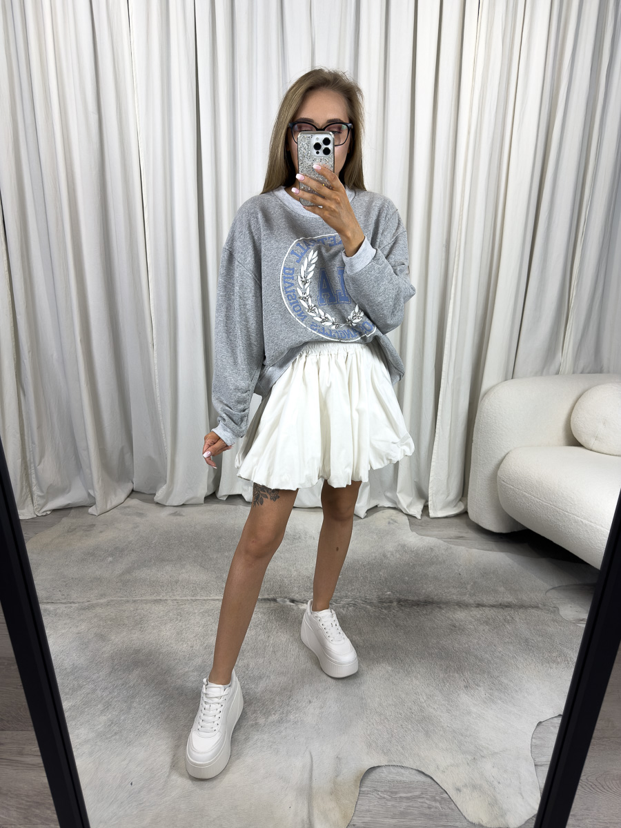 Bluza oversize College szara z nadrukiem college, stylizacja z białą spódnicą i białymi sneakersami, zdjęcie produktowe na tle jasnych zasłon.