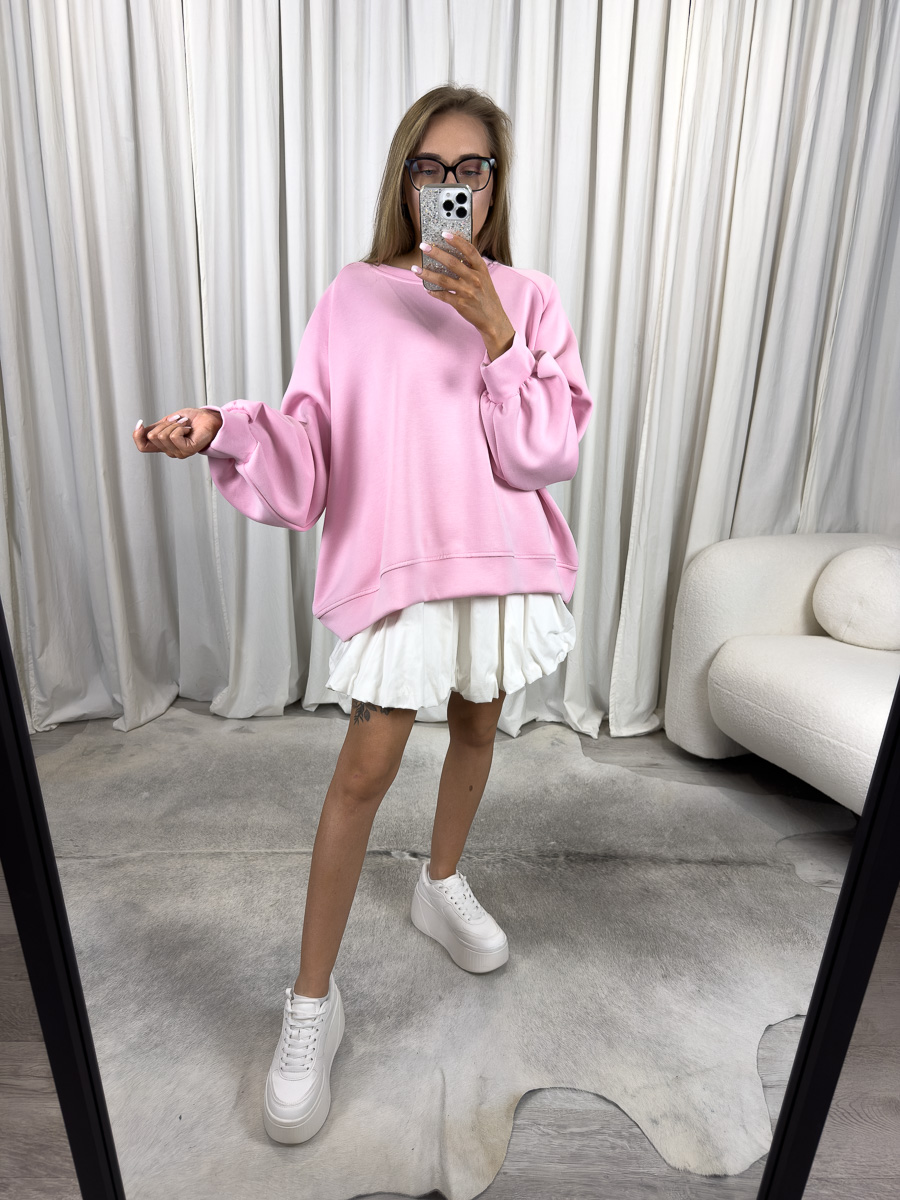 Modelka w pastelowej różowej bluzie oversize Casual z białą plisowaną spódnicą i różową torebką na złotym łańcuszku – stylizacja miejska z dziewczęcym akcentem.