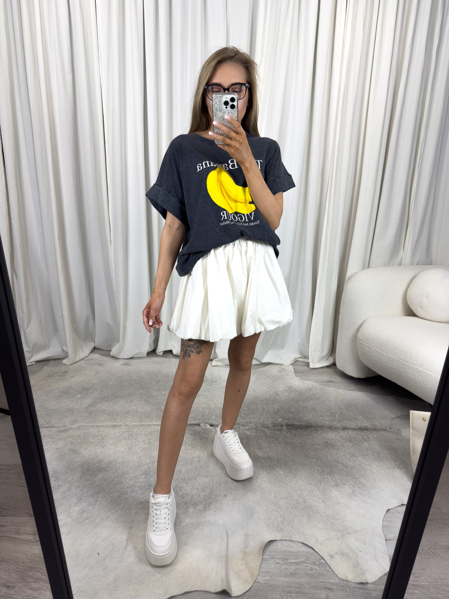 Modelka w dekatyzowanym T-shircie z nadrukiem banana i białej spódniczce – stylizacja z przymrużeniem oka, casualowa i dziewczęca.