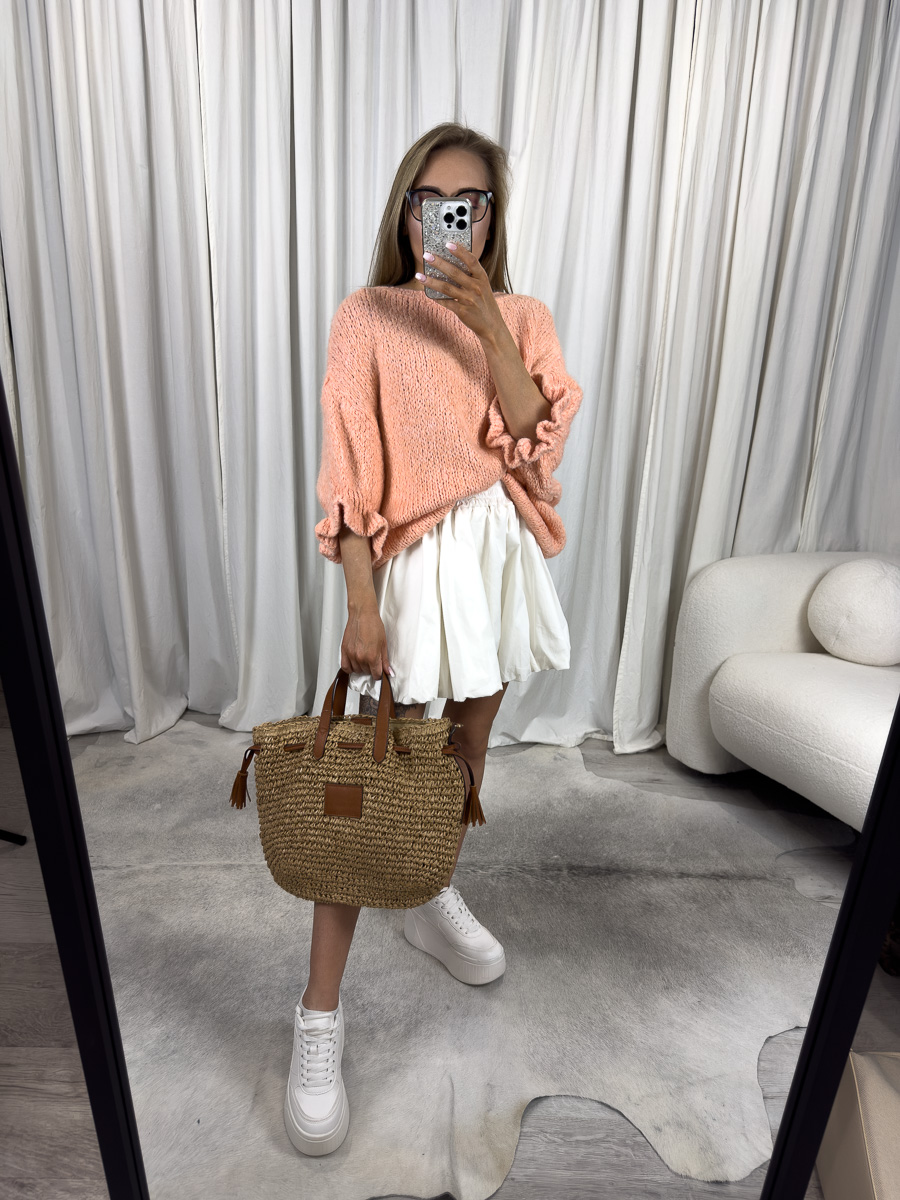Kobieta w swetrze pastelowym Flounce pomarańczowym z falbanką i białą spódnicą, z plecioną torbą – stylizacja casual boho.