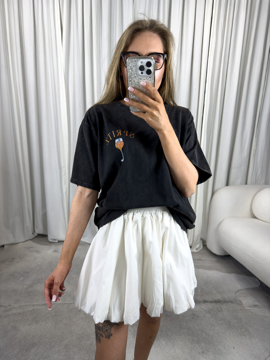 Modelka ubrana w dekatyzowany T-shirt z nadrukiem Spiritz Club, w białą mini spódniczkę oraz białe wysokie sneakersy.