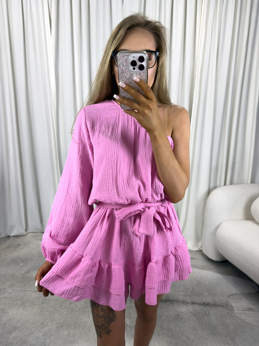 muslinowy-kombinezon-girlish-rozowy Modelka w muślinowym kombinezonie Girlish z asymetrycznym ramieniem, falbankami i wiązaniem w talii. Zdjęcie produktowe sklepu internetowego z odzieżą damską.